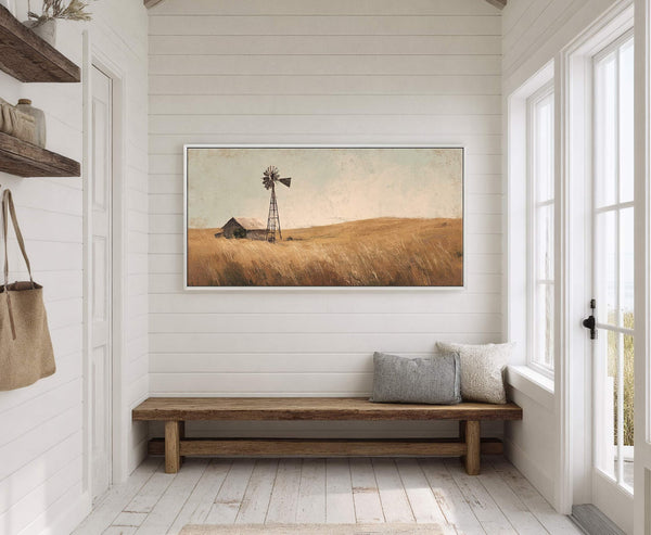 This rustic landscape painting print depicts a windmill and barn in a golden field with a pale sky, framed in white and hung on a white plank wall above a.