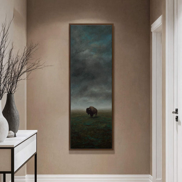 This vertical canvas print depicts a lone bison in a misty field under a dramatic, dark sky, rendered in a moody, atmospheric style with a desaturated palette, framed in dark.