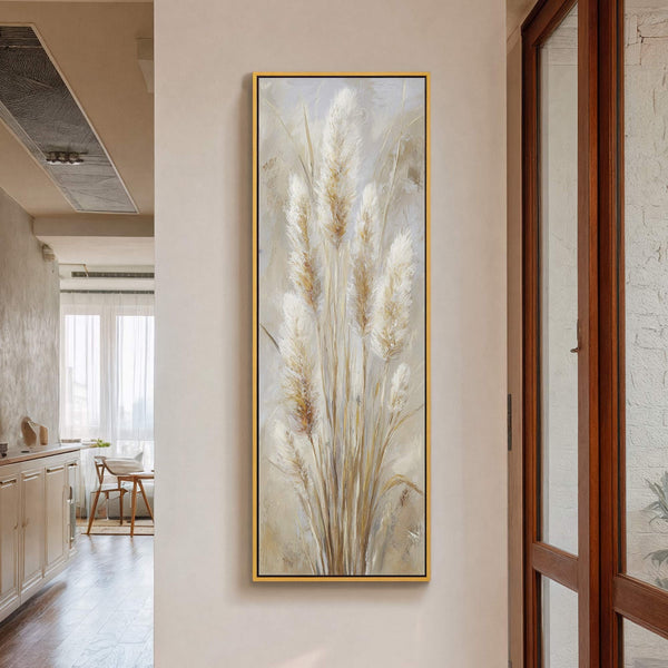This vertical canvas wall art features a painterly depiction of pampas grass in a neutral palette, framed in gold, hanging on a light-toned wall with a wooden door visible to.