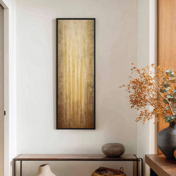 This vertical, abstract art print features a textured palette of gold and brown hues, presented in a black floater frame, and hangs on a white wall above a wooden console.