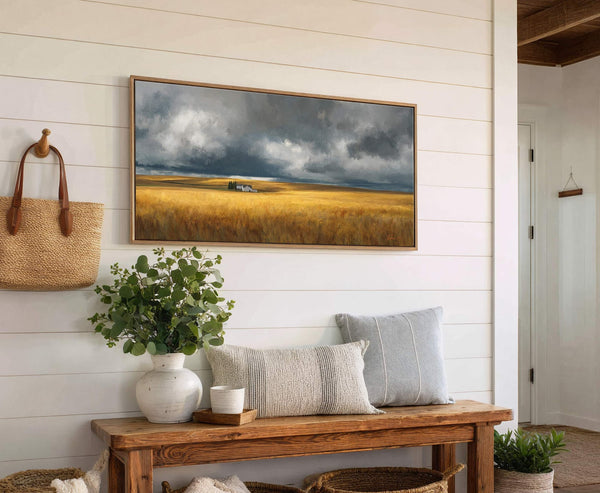 This horizontal canvas wall art features a stormy sky over a golden field with distant white buildings, rendered in a moody impressionistic style. It is displayed on a white shiplap.