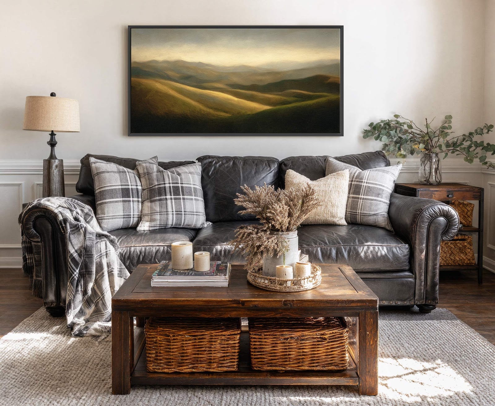 This horizontal canvas wall art features a landscape painting with rolling hills illuminated by warm, golden light. The artwork is framed in black and hangs above a dark leather sofa.