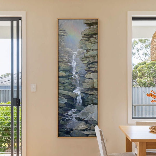 This vertical canvas wall art depicts a realistic waterfall cascading through rocky terrain with a subtle rainbow in the background. The earthy palette features grays and greens. It is displayed.