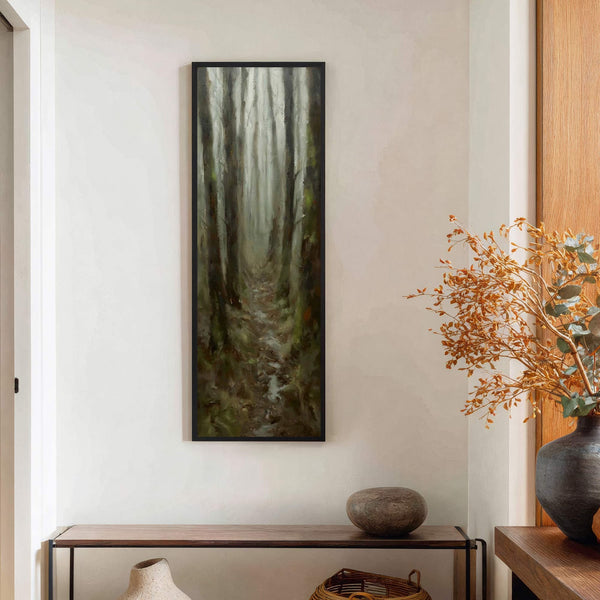 This vertical canvas wall art depicts a misty forest path with a muted green and brown palette, in an impressionistic style. It hangs on a white wall above a wooden.