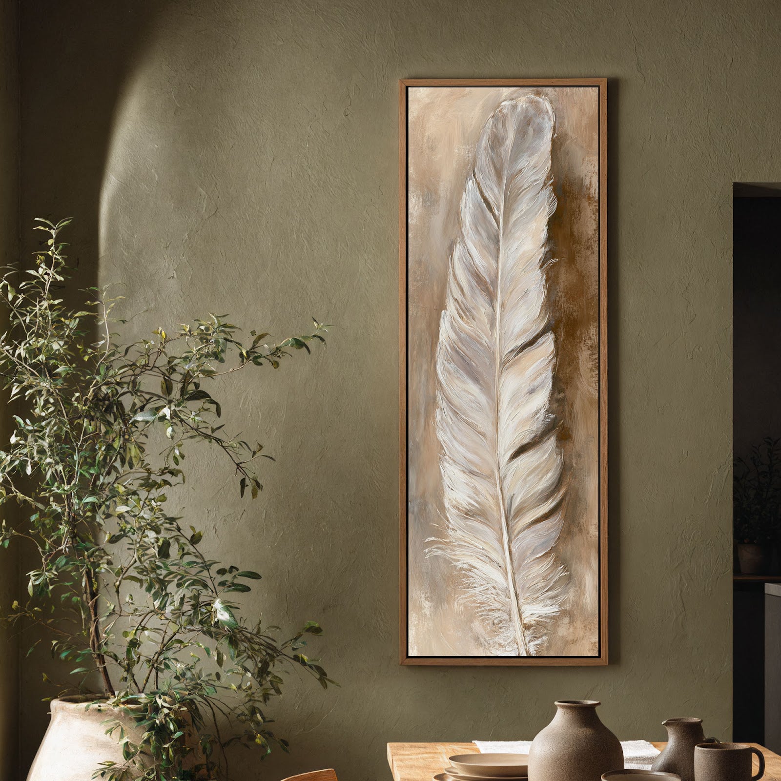 This vertical art print features a textured white feather in earthy tones, showcased in an oak floater frame on a sage green plaster living room wall beside a large plant.