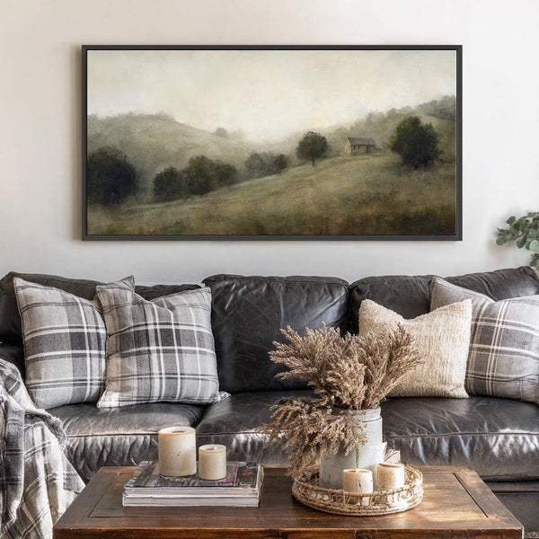 This horizontal canvas wall art features a misty landscape with rolling hills and trees, rendered in a muted green and gray palette. It hangs above a dark leather sofa in.
