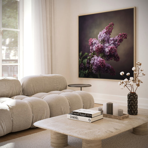 Large natural framed opulent lilac botanical art print featuring vibrant purple and fuchsia lilac clusters against a dark, moody background, displayed on a white wall above a beige sofa and natural stone coffee table in a bright, modern living room.