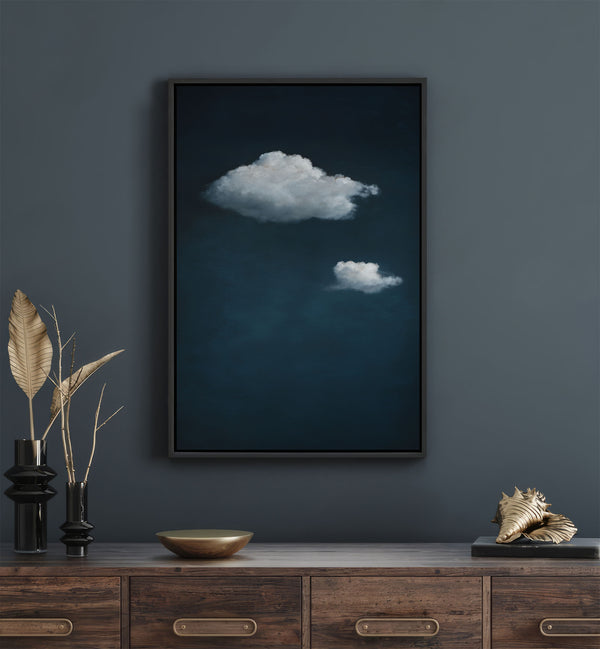 Black-framed minimalist art print featuring two white clouds floating against a dark blue textured sky, displayed above a wooden console table with decorative objects.