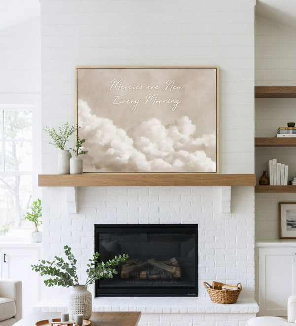 Large natural framed inspirational art print featuring soft, painterly white clouds against a warm beige sky with the elegant script text 'Mercies are New Every Morning', displayed above a white brick fireplace with a natural wood mantel in a bright, modern living room.