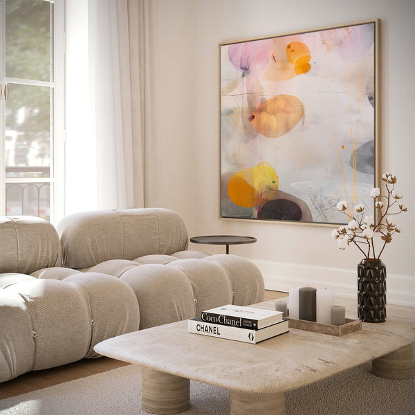Large natural framed abstract art print featuring layered, fluid shapes in soft pink, orange, yellow, and grey against a light textured background, displayed above a beige modular sofa and natural stone coffee table in a bright living room