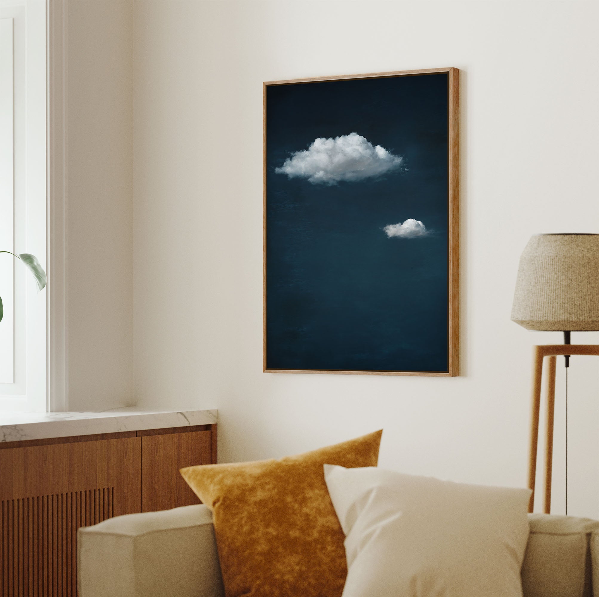 Pecan-framed minimalist art print featuring two soft white clouds floating against a deep, dark blue sky with subtle texture, displayed on a light wall above a cream sofa in a bright living room with a window.