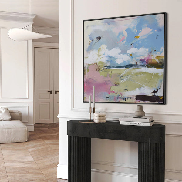 Black-framed abstract painting featuring soft, expressive brushstrokes in light blues, pinks, whites, and greens, resembling a dreamy sky over a field, displayed above a black console table in a minimalist white entryway