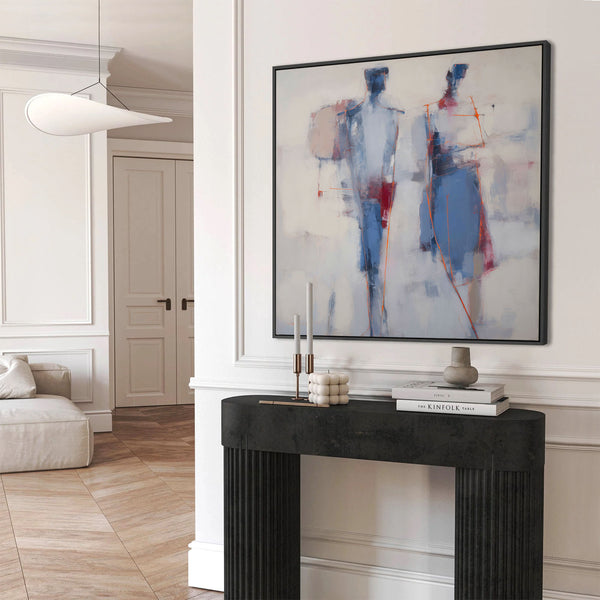 Large black-framed abstract art print depicting two painterly, elongated figures in shades of blue, white, red, and orange, against a light textured background, displayed above a black console table in a minimalist white entryway.