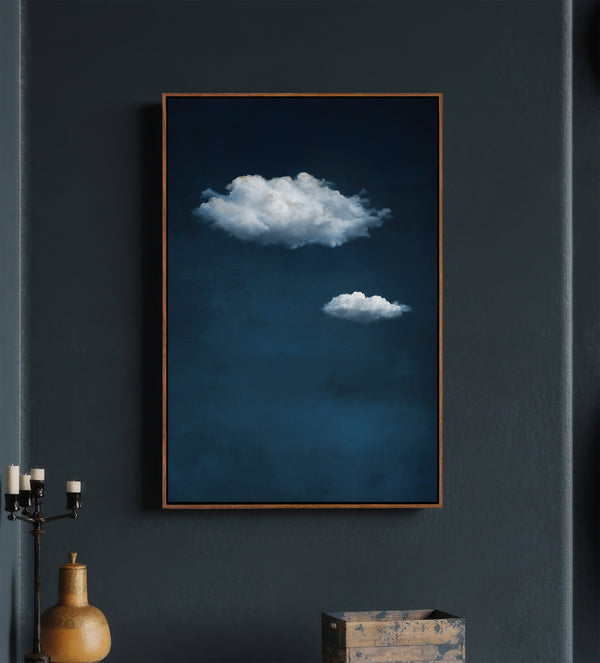 Pecan framed minimalist art print featuring two soft white clouds floating against a deep, dark blue textured sky, displayed on a dark blue wall above a console table