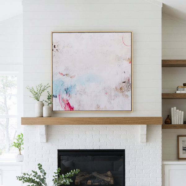 Large natural framed abstract art print featuring soft blush pink, serene blue, and subtle black painterly strokes and organic textures on a predominantly white and off-white background, displayed above a white brick fireplace with a natural wood mantel in a bright, modern living room with white shiplap walls.