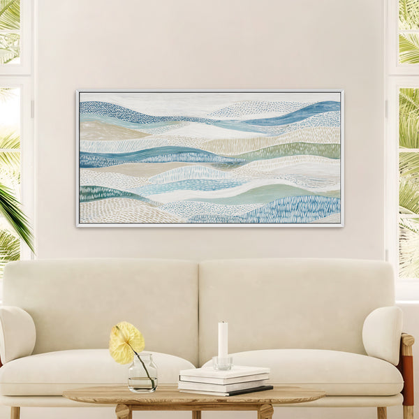 Large white-framed abstract art print featuring layered, textured patterns in shades of blue, green, and beige, resembling rolling waves or hills, displayed above a cream-colored sofa and wooden coffee table in a bright, modern living room with large windows