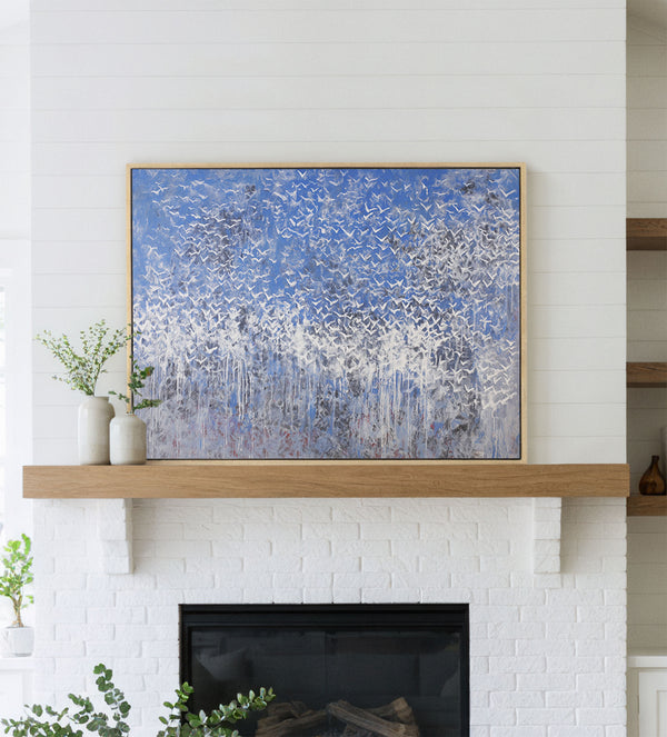 Large framed giclee art print featuring an abstract flock of white birds against a blue sky, displayed above a white brick fireplace with a wooden mantel in a modern living room