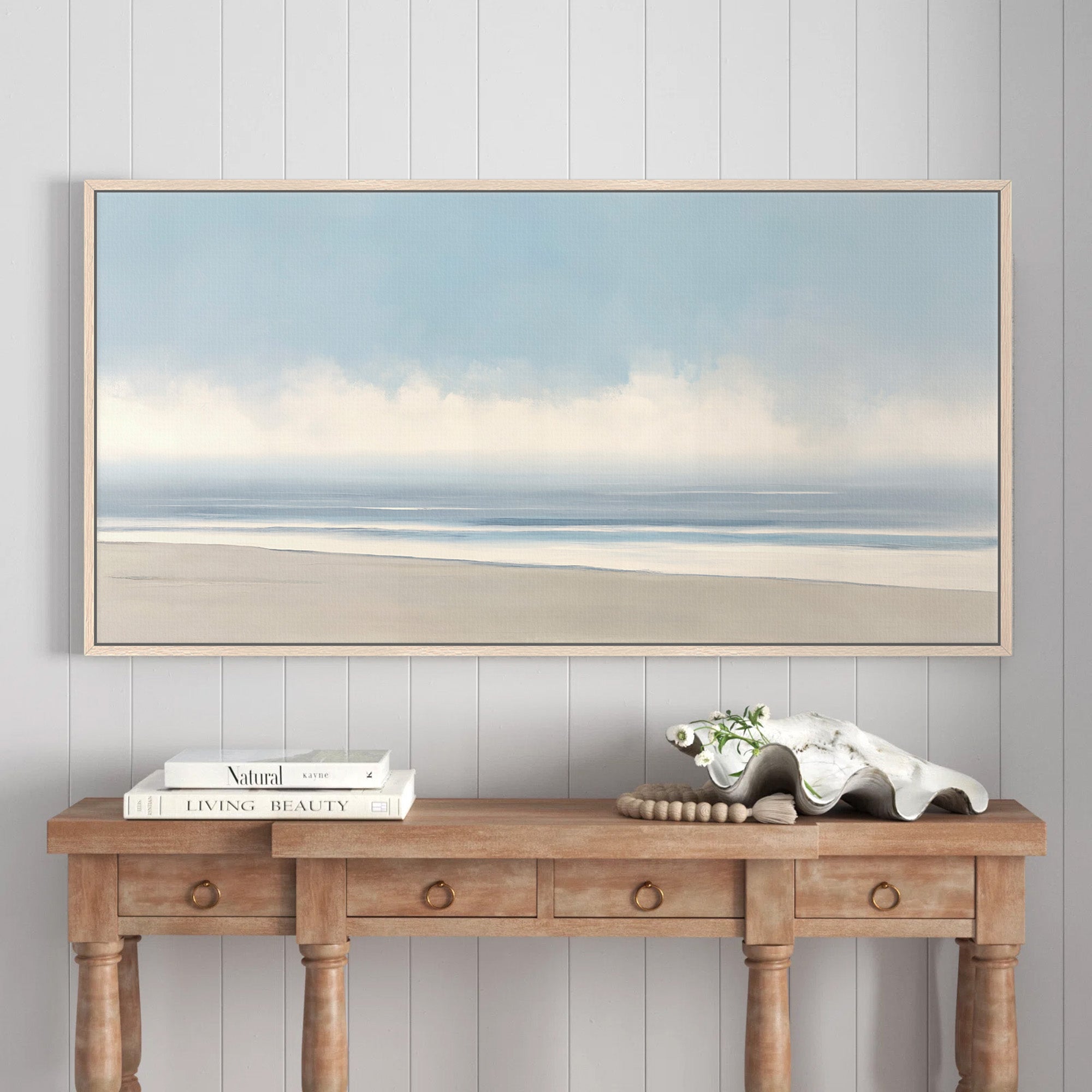 Framed minimalist coastal landscape canvas print featuring a serene beach, ocean, and sky in soft blue and sand tones, hanging above a wooden console table.