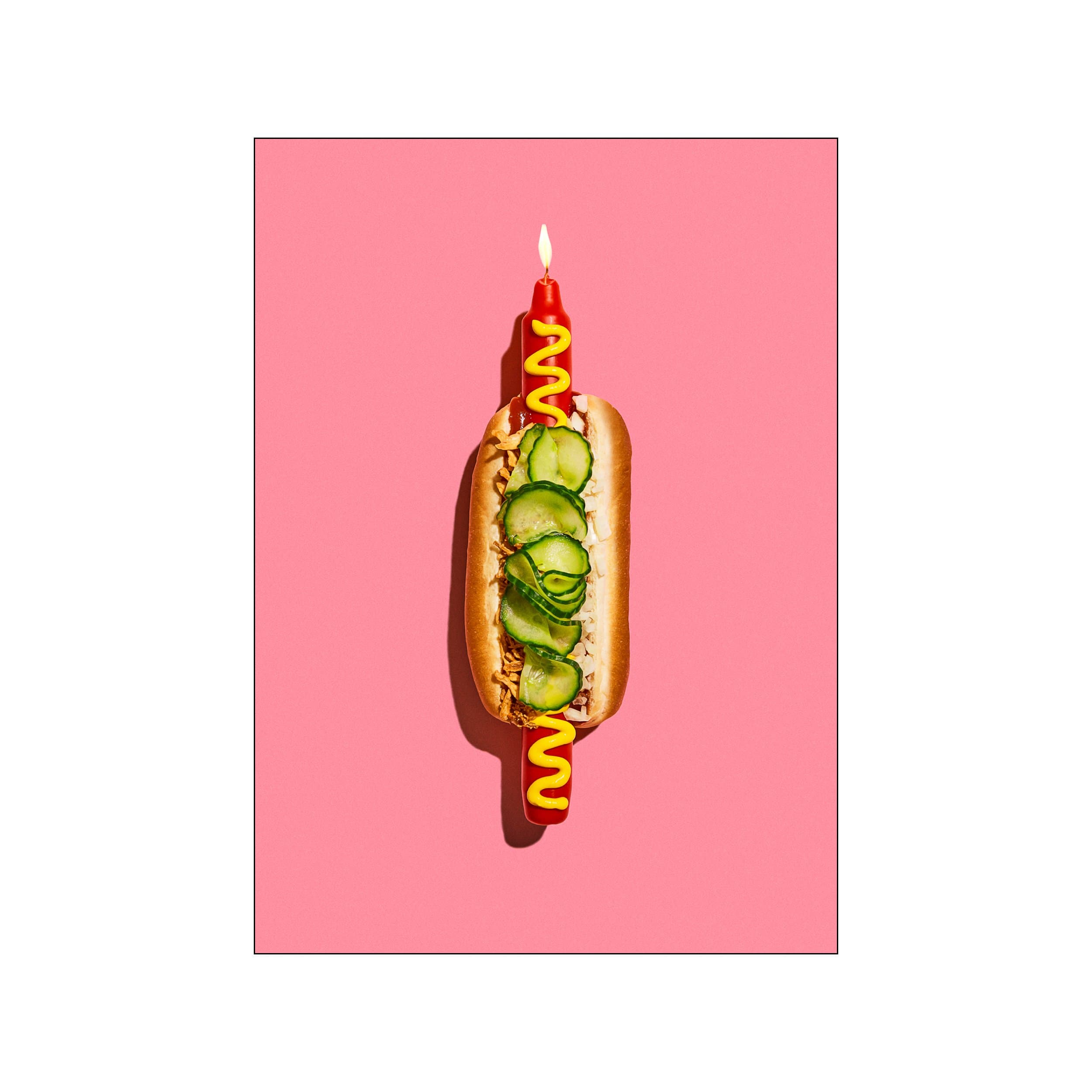 Surreal hot dog topped with a candle, mustard, and cucumber slices on a pink background