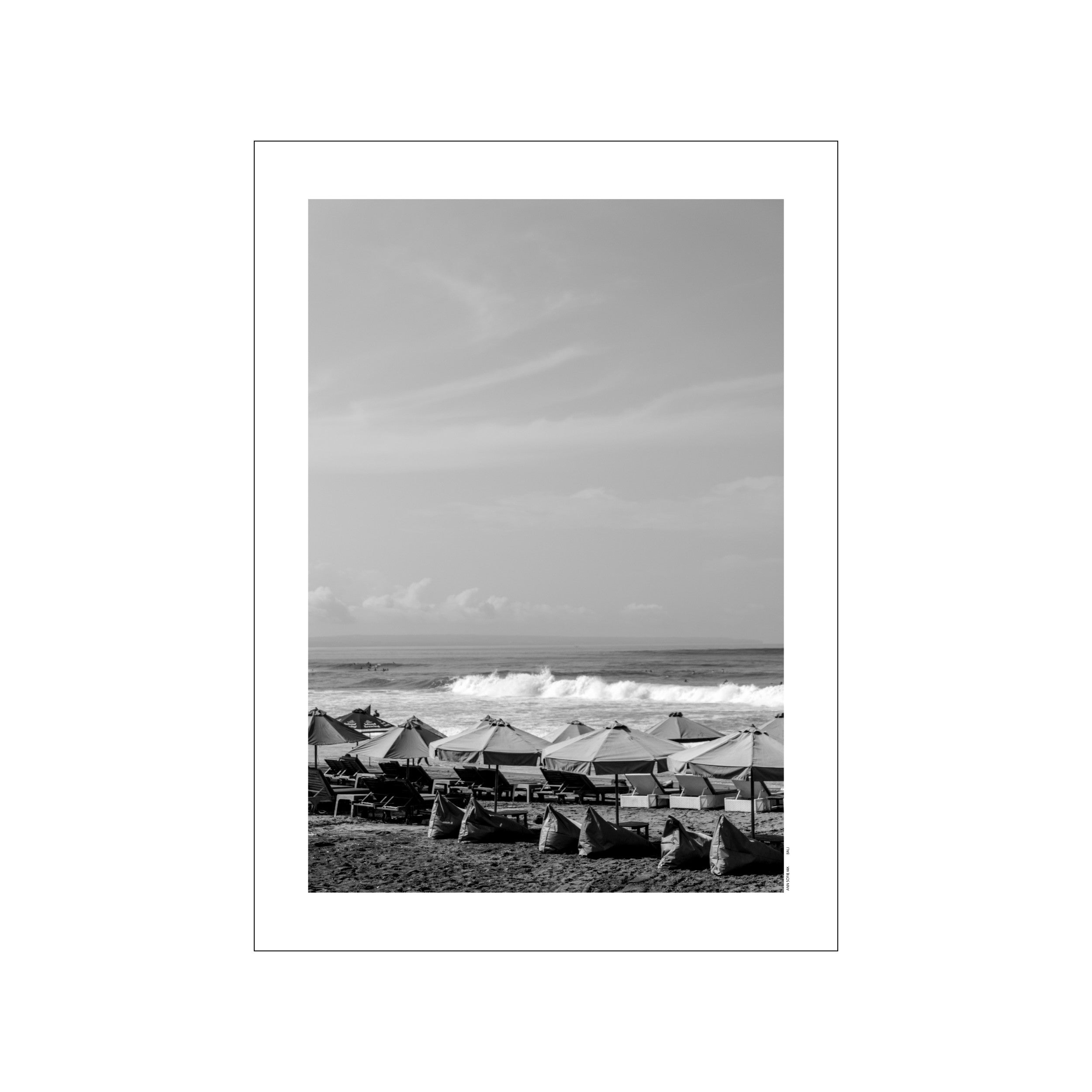 A black and white photograph of Canggu Beach with rows of beach umbrellas and chairs on the sand, and gentle waves rolling into the shore under a cloudy sky.