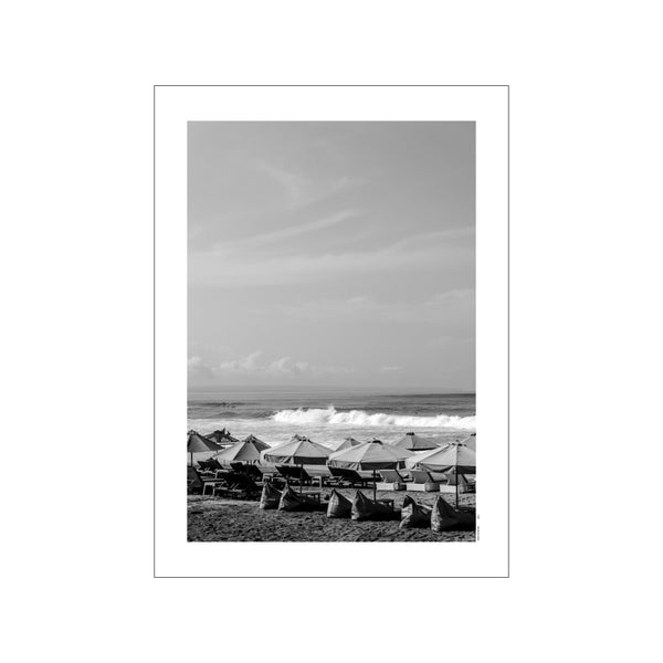 A black and white photograph of Canggu Beach with rows of beach umbrellas and chairs on the sand, and gentle waves rolling into the shore under a cloudy sky.