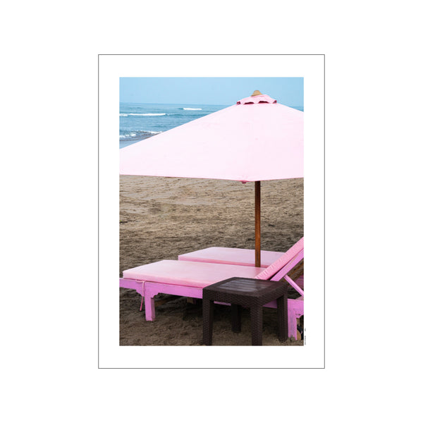 Two plush pink sunbeds and a large pale pink umbrella sit on a sandy beach, with gentle blue waves in the background. The mood is peaceful and serene.
