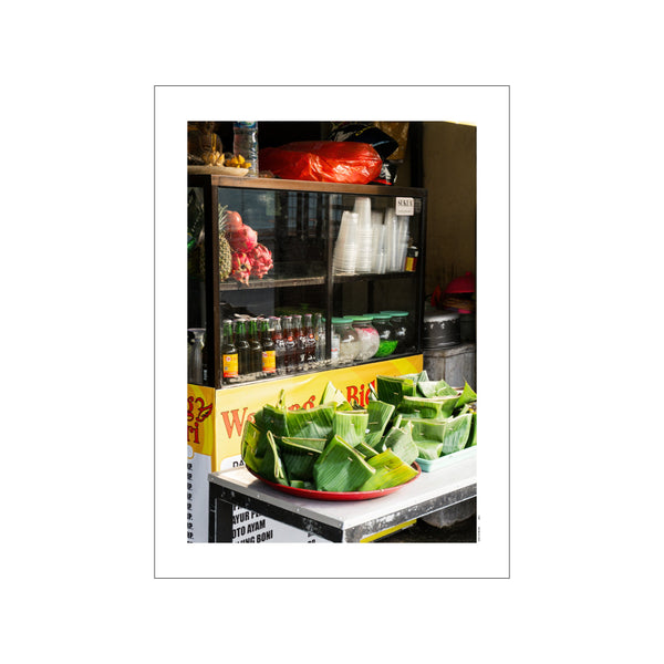 A close-up of food parcels wrapped in bright green banana leaves, displayed on a red tray in front of a glass-fronted stall filled with drinks and fruit.