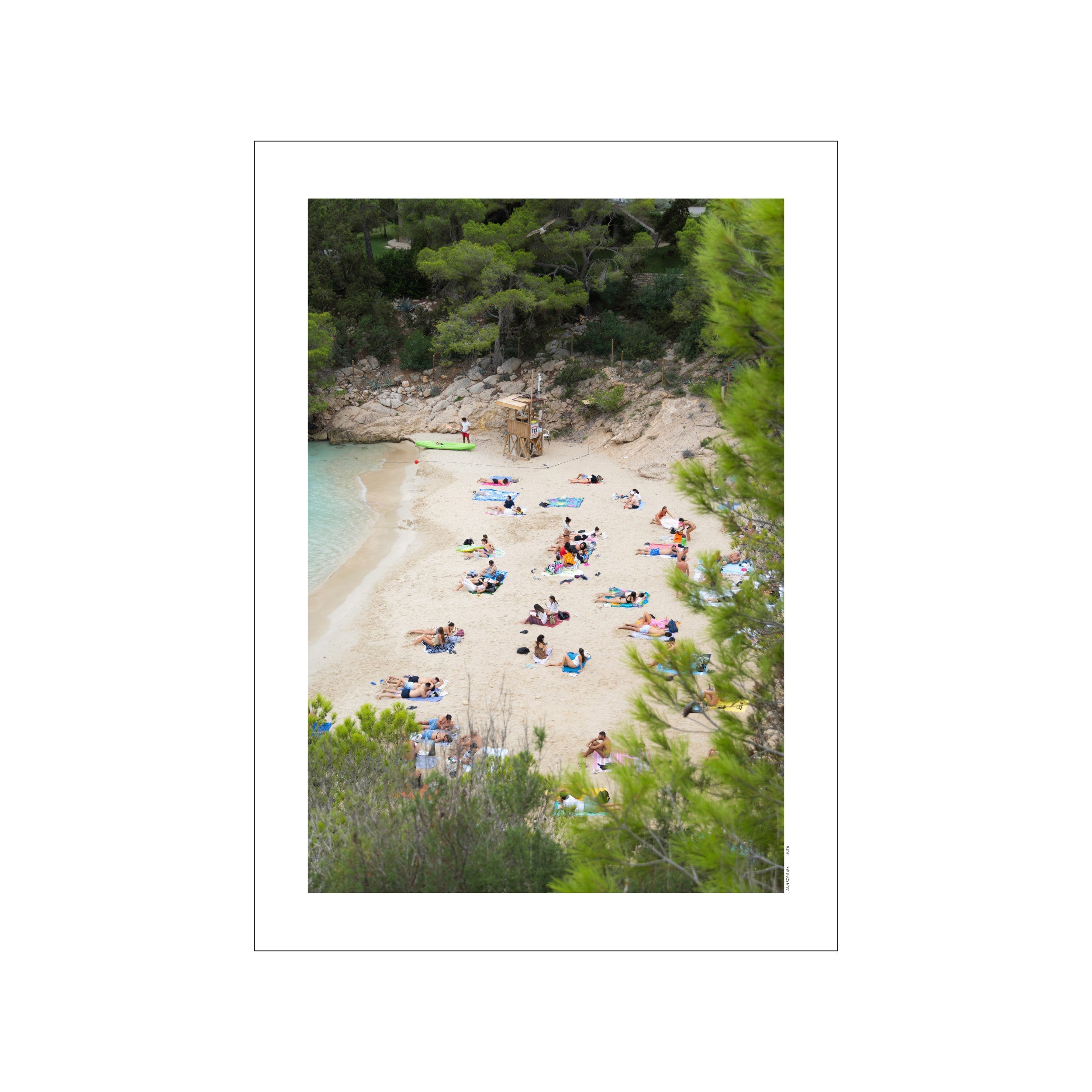 An aerial view of a sandy cove in Ibiza, dotted with sunbathers and framed by lush green pine trees, with clear turquoise water lapping the shore.