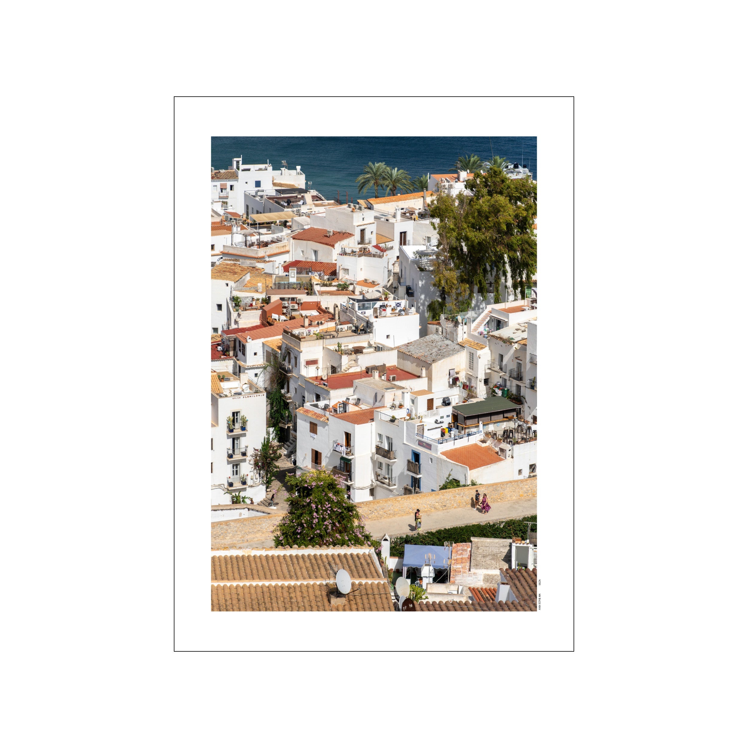 An aerial view captures the charming streets and whitewashed architecture of Ibiza Town, with terracotta roofs and the blue Mediterranean Sea in the background, evoking a serene coastal vibe.