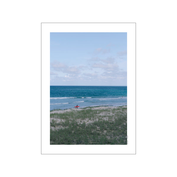 A person in silhouette walks along a sandy dune toward the turquoise ocean holding a bright red surfboard, under a pale blue sky with wispy clouds.