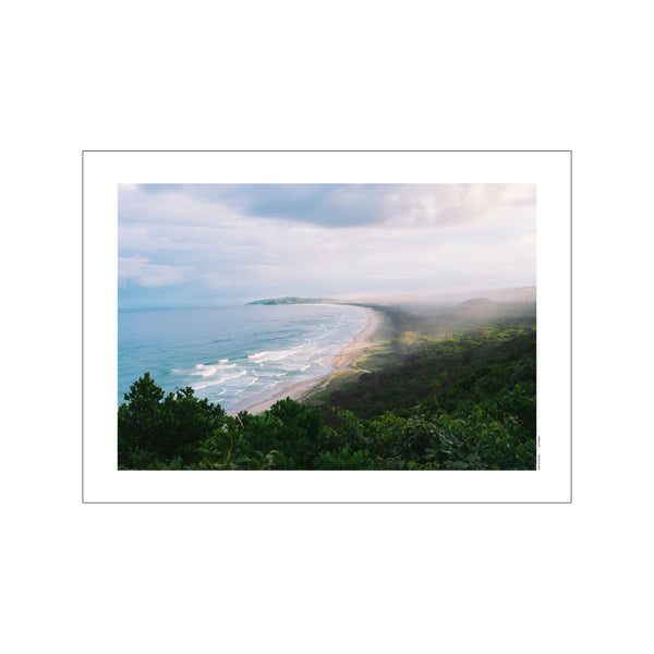 An elevated view of a curving beach and turquoise ocean waves under a soft, misty sky, framed by lush green trees in the foreground. The atmosphere is peaceful and serene.